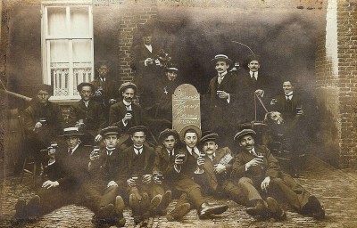 Gezelschap jongeheren met een glaasje bier rondom een bord 'Heeswijk Hoe wij kermis vieren'. De foto is gemaakt bij café Van Oorschot of het café tegenover Cunera. Heeswijk ca. 1922