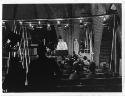 Werkfoto van opnames film Zonnelied, Etten 1938. Filmscene van een preek van de pastoor van Overa over het belang van goede gezondheidszorg (foto: fotograaf onbekend, collectie Museum voor Verpleegkunde Culemborg)